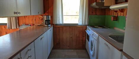 Ett kompakt kök med träväggar, en tvättmaskin, en kaffebryggare och ett fönster med gardiner.