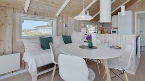 A cozy cabin interior with a round dining table, white chairs, a sofa with green cushions, and a large window offering a view of a building outside. - "Betti" - 300m from the sea by Interhome (Rømø)