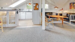 Amplia sala de estar con techo alto, una mesa de comedor y un sofá. Se ve una cocina en el fondo con armarios blancos y un aparador de vidrio.