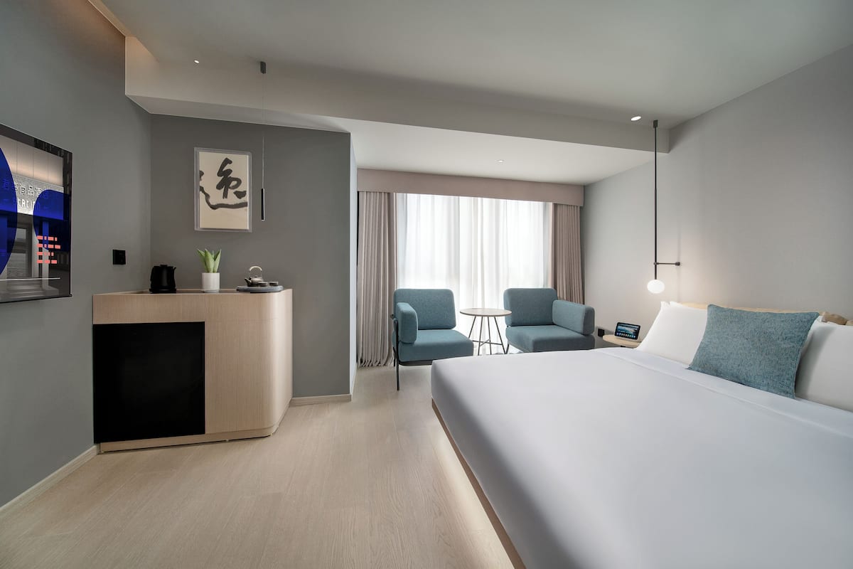 Ein modernes Hotelzimmer mit einem großen Bett, einem Schreibtisch mit Pflanze, einem kleinen Tisch, zwei Stühlen und einem großen Fenster mit Vorhängen.