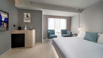 A modern hotel room with a large bed, a desk with a plant, a small table, two chairs, and a large window with curtains.