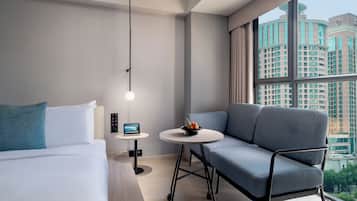 A modern hotel room with a bed, a small round table with a fruit bowl, a laptop, and a seating area with a chair.