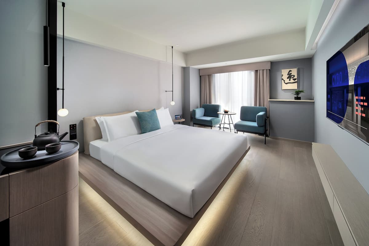 Ein modernes Hotelzimmer mit einem großen Bett, Nachttischen, einem Schreibtisch mit Wasserkocher und einer Sitzecke mit Stühlen und einem kleinen Tisch.