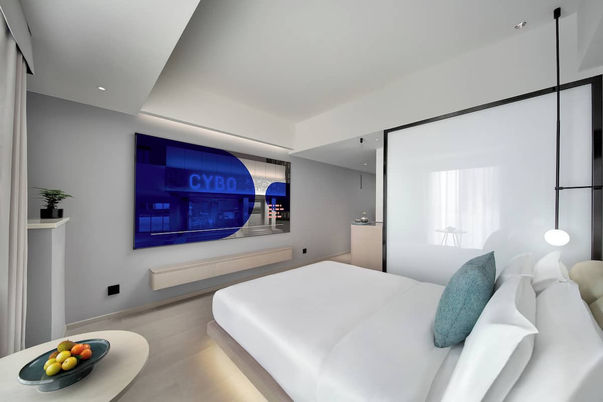 Ein modernes Schlafzimmer mit einem großen Bett, einem flachbildfernseher an der Wand und einer Obstschale auf einem Nachttisch.