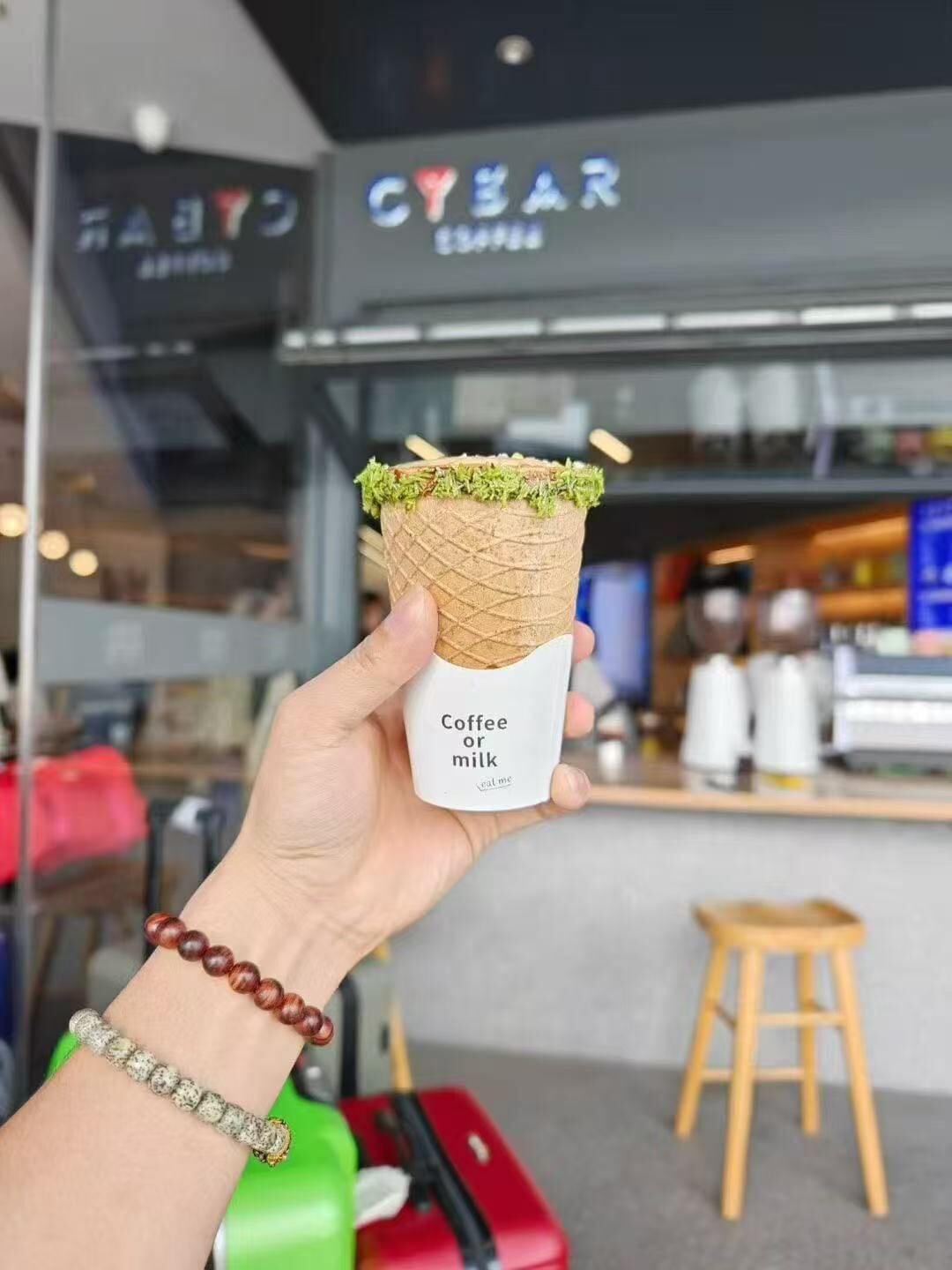 A hand holding a coffee or milk cone with a label, in front of a coffee shop named CYBAR.