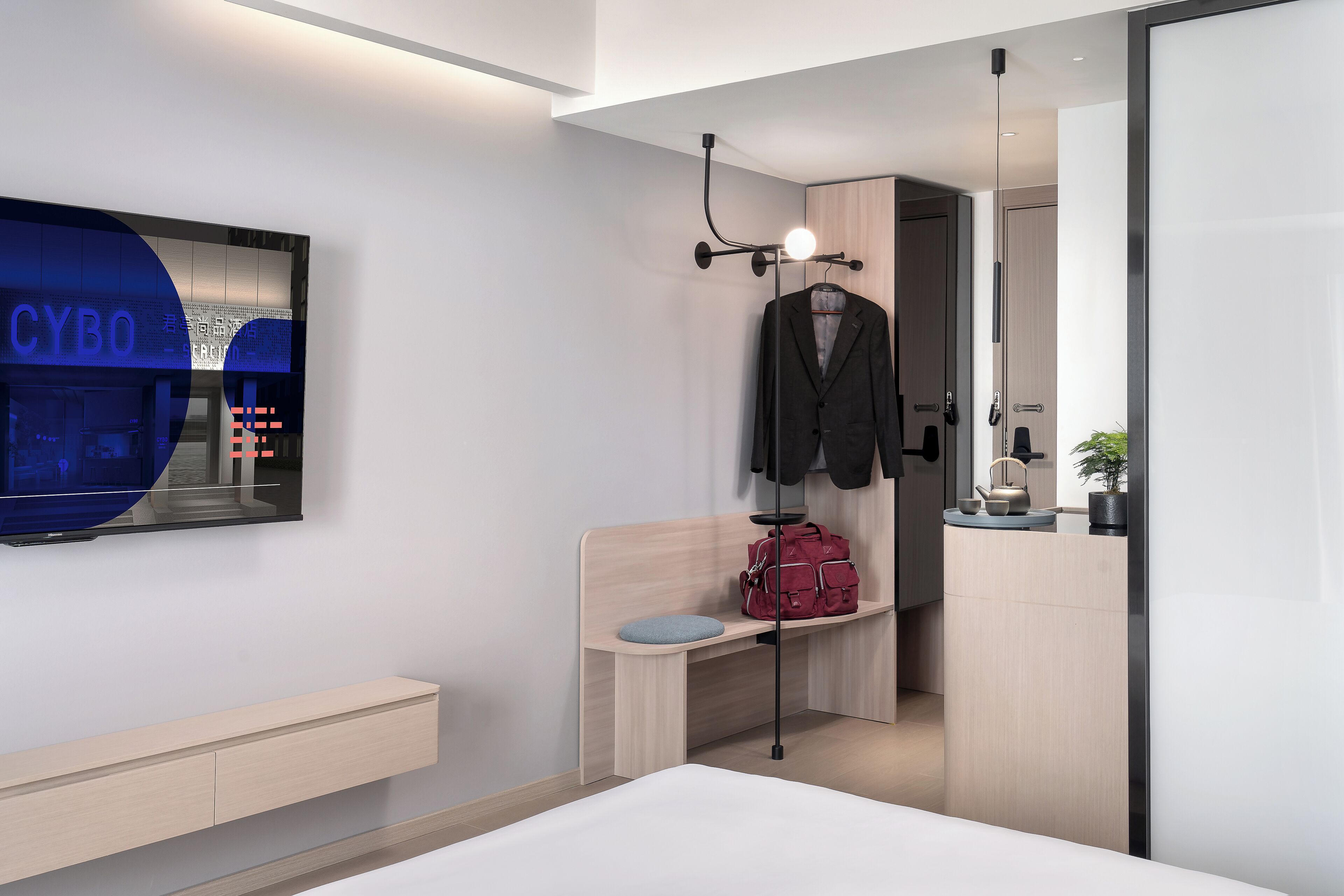 Ein modernes Hotelzimmer mit Bett, einer Sitzbank, einem hängenden Kleiderständer, einer roten Tasche und einem an der Wand montierten Fernseher, der 'CYBO' zeigt.