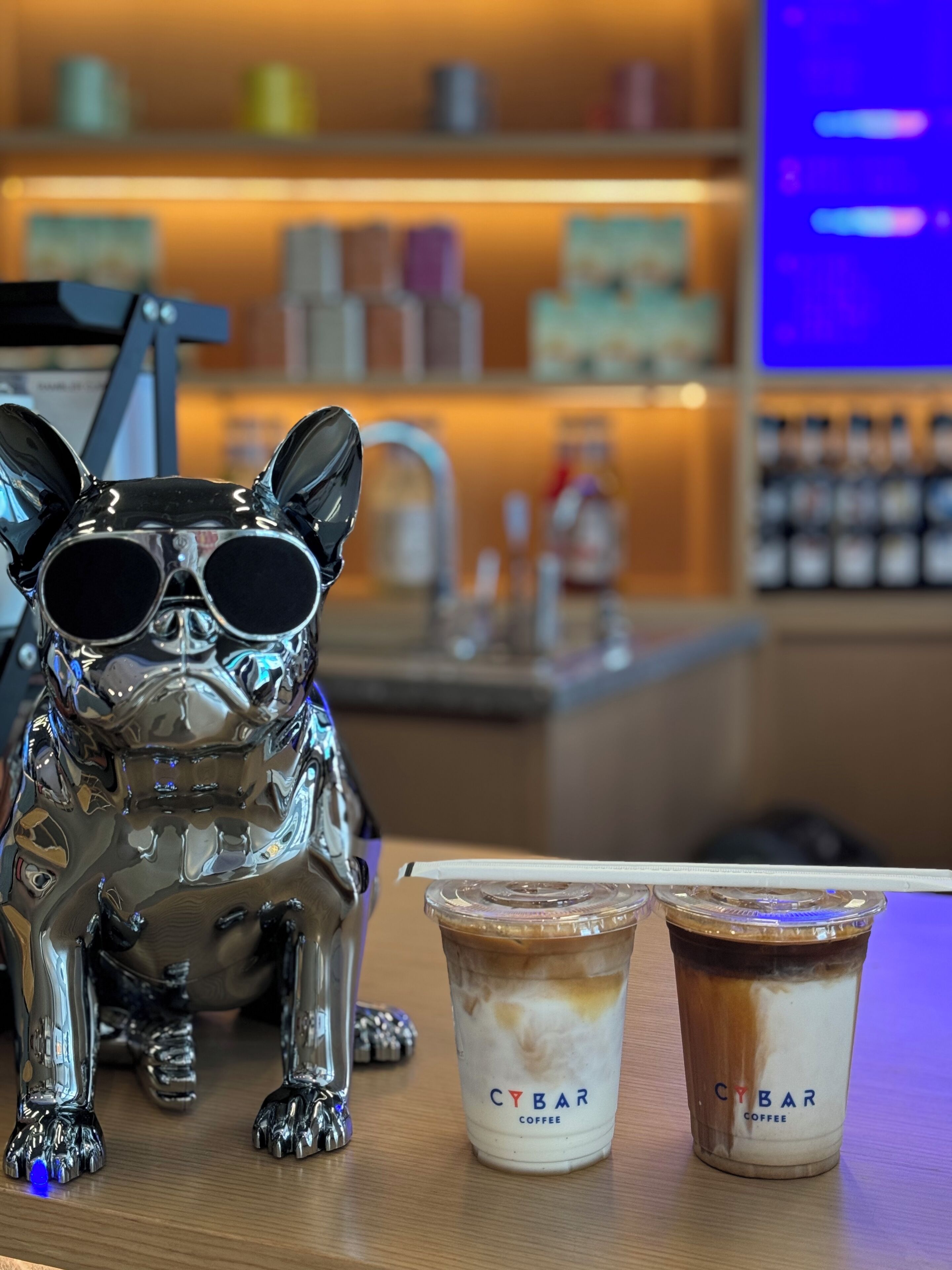 A metallic dog figurine with sunglasses, two Cibar Coffee cups, and a blurred background with shelves.