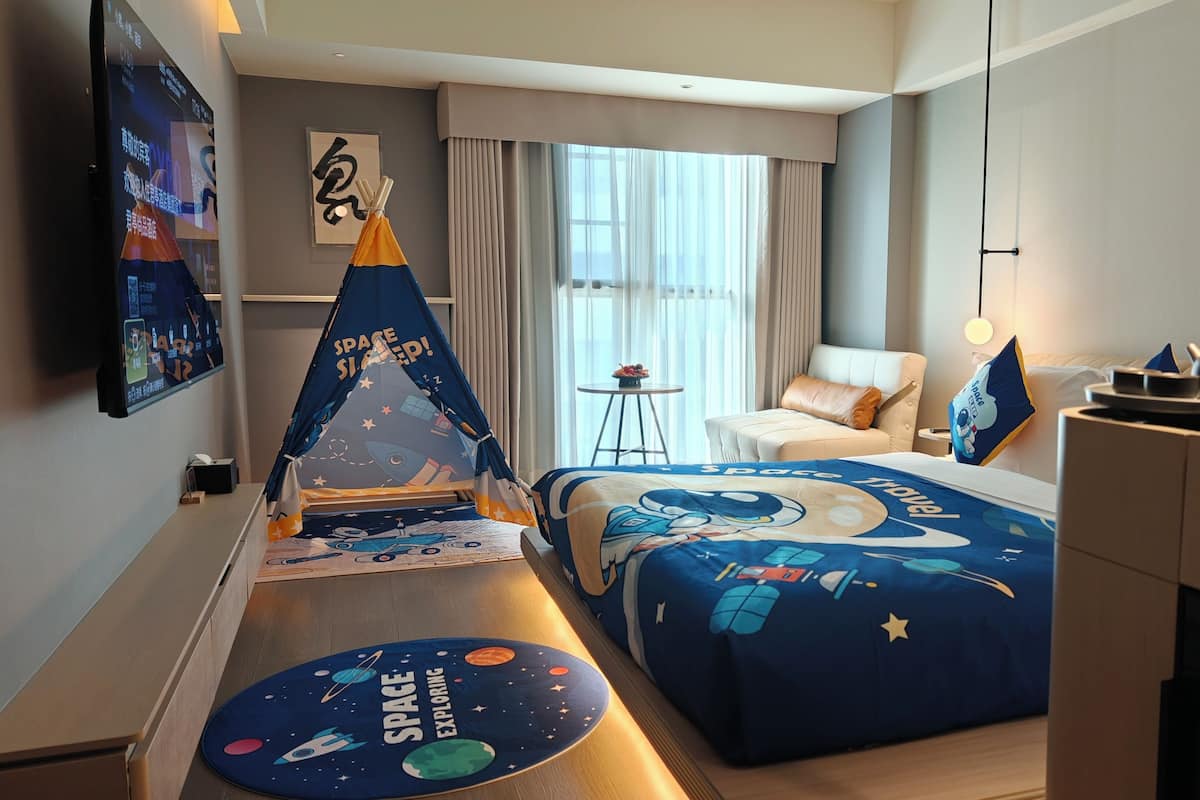 Ein Hotelzimmer mit Bettwäsche zum Thema Weltraum, einer zeltartigen Dekoration und einem an der Wand montierten Fernseher, der eine Benutzeroberfläche mit Weltraummotiv zeigt.