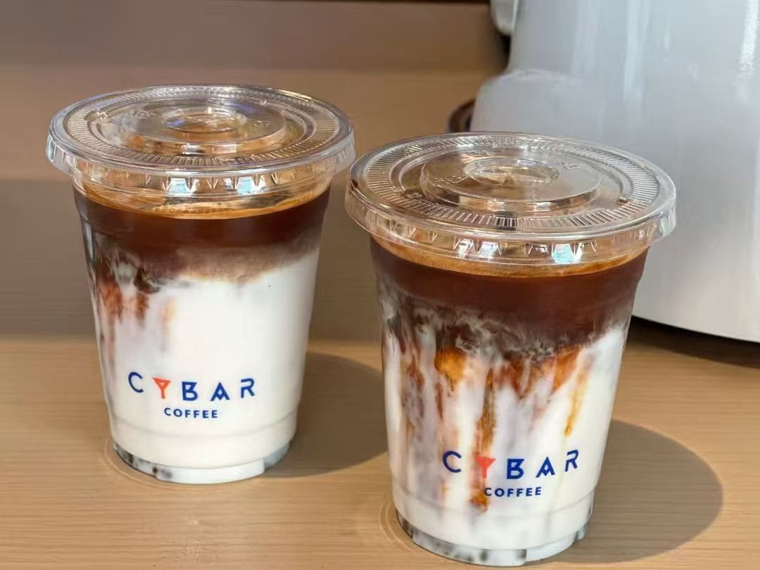 Dos tazas de CYBAR COFFEE con tapas sobre una superficie de madera.