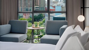 Un hotel moderno con dos sillones grises, una pequeña mesa redonda con una botella y un vaso, y vistas de la ciudad a través de grandes ventanales.