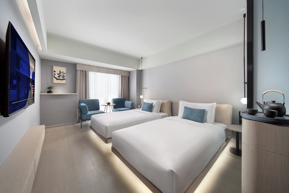 Ein modernes Hotelzimmer mit zwei Betten, einem Fernseher, einem Schreibtisch und einem kleinen Tisch mit einer Pflanze.