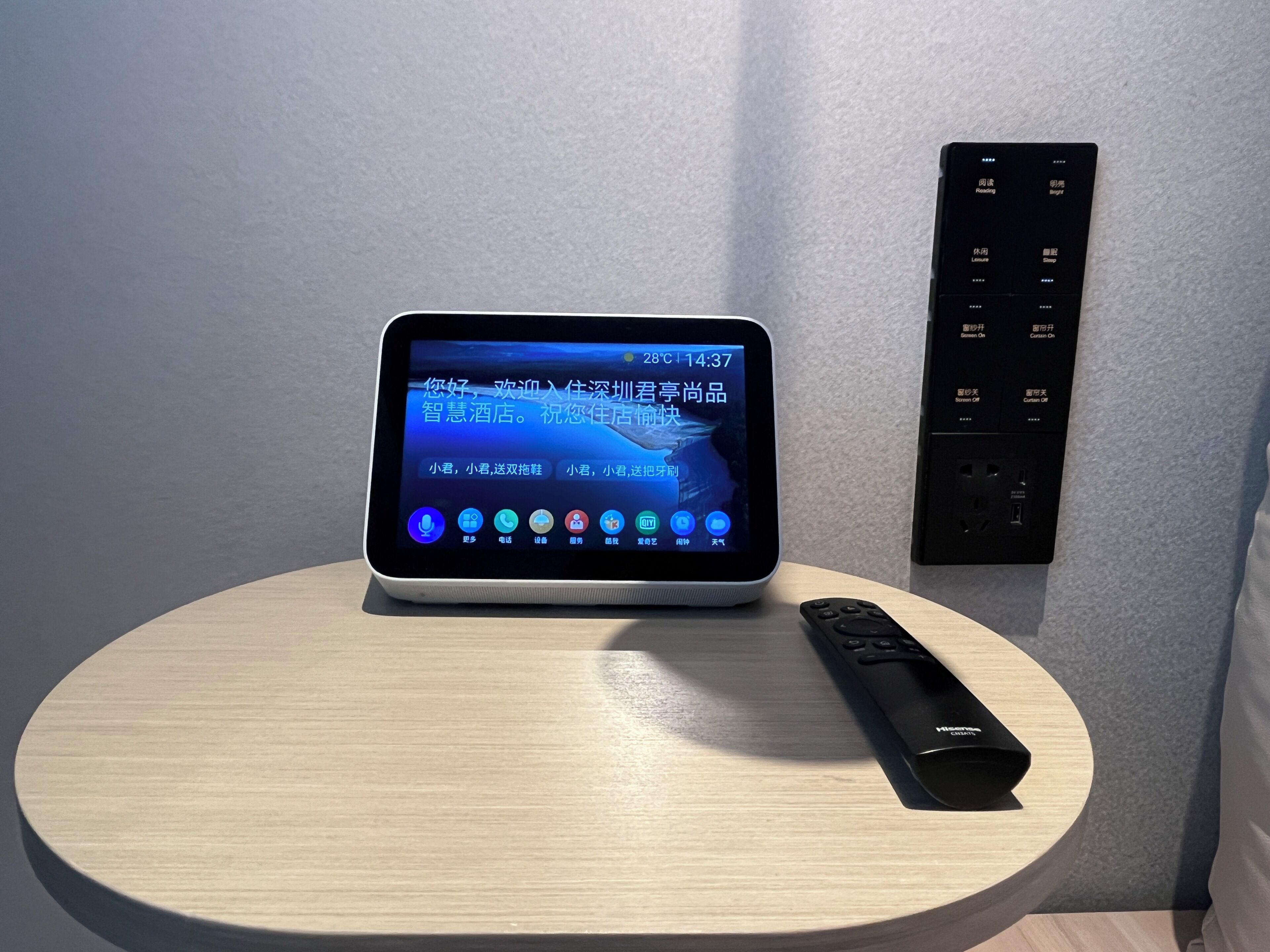 Une tablette posée sur une table ronde affiche un message d’accueil de l’hôtel en chinois. Une télécommande se trouve à côté, et un panneau de commande fixé au mur présente divers boutons.