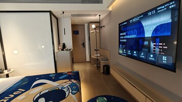 A hotel room with a large digital display on the wall, a bed with a space-themed bedspread, a bedside table with a bottle and a cup, and a small trash bin.