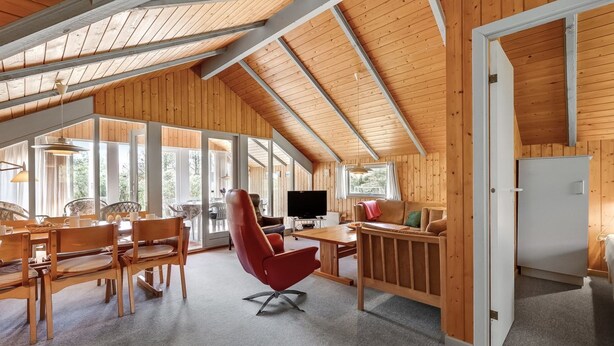 Amplia habitación tipo loft con techo alto, vigas de madera, zona de comedor con mesa y sillas, una silla roja, sofá y televisor.