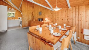 Interior de una cabaña de madera con una mesa de comedor preparada para cuatro comensales, una zona de cocina con electrodomésticos modernos y un gran ventanal que proporciona luz natural.