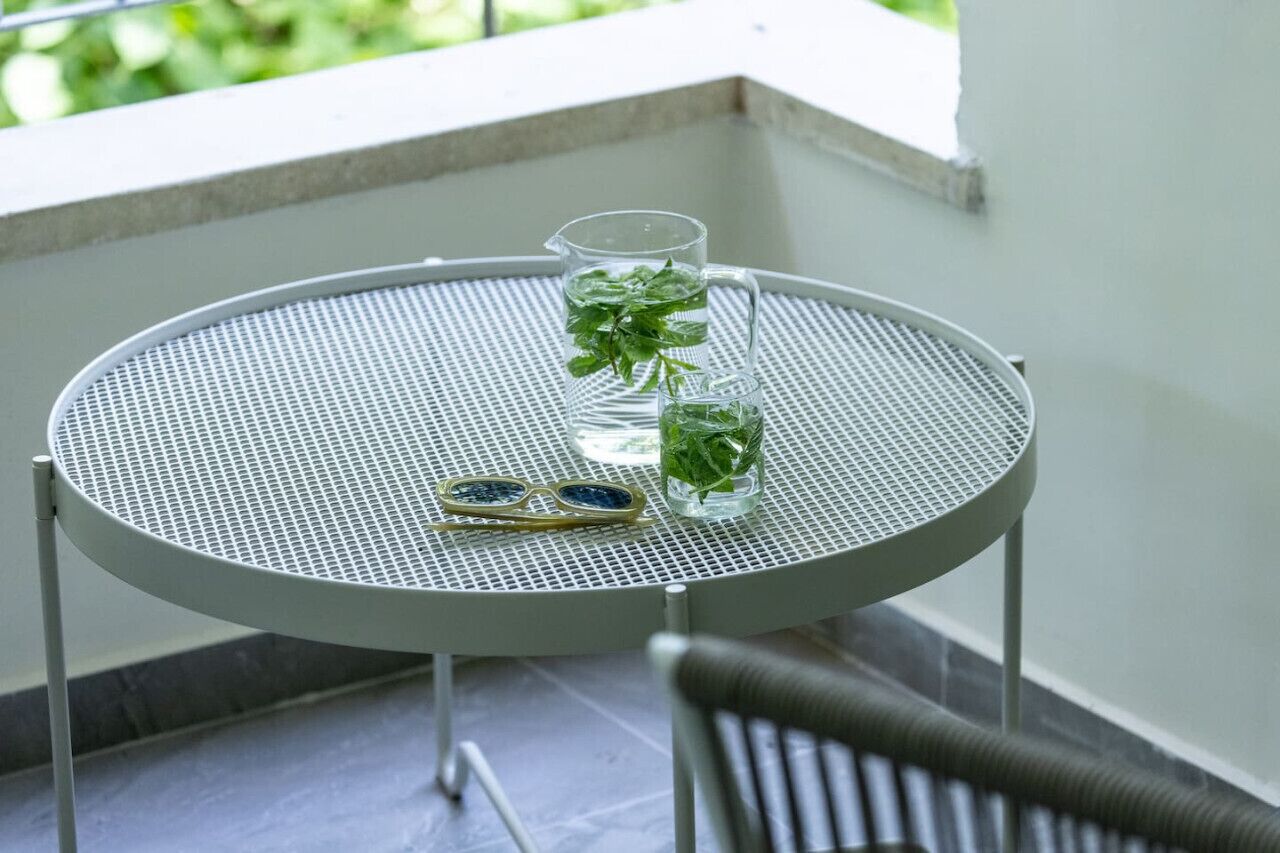 Una mesa metálica redonda con superficie de malla, una jarrón con agua, un vaso con bebida y un par de gafas de sol sobre la mesa.