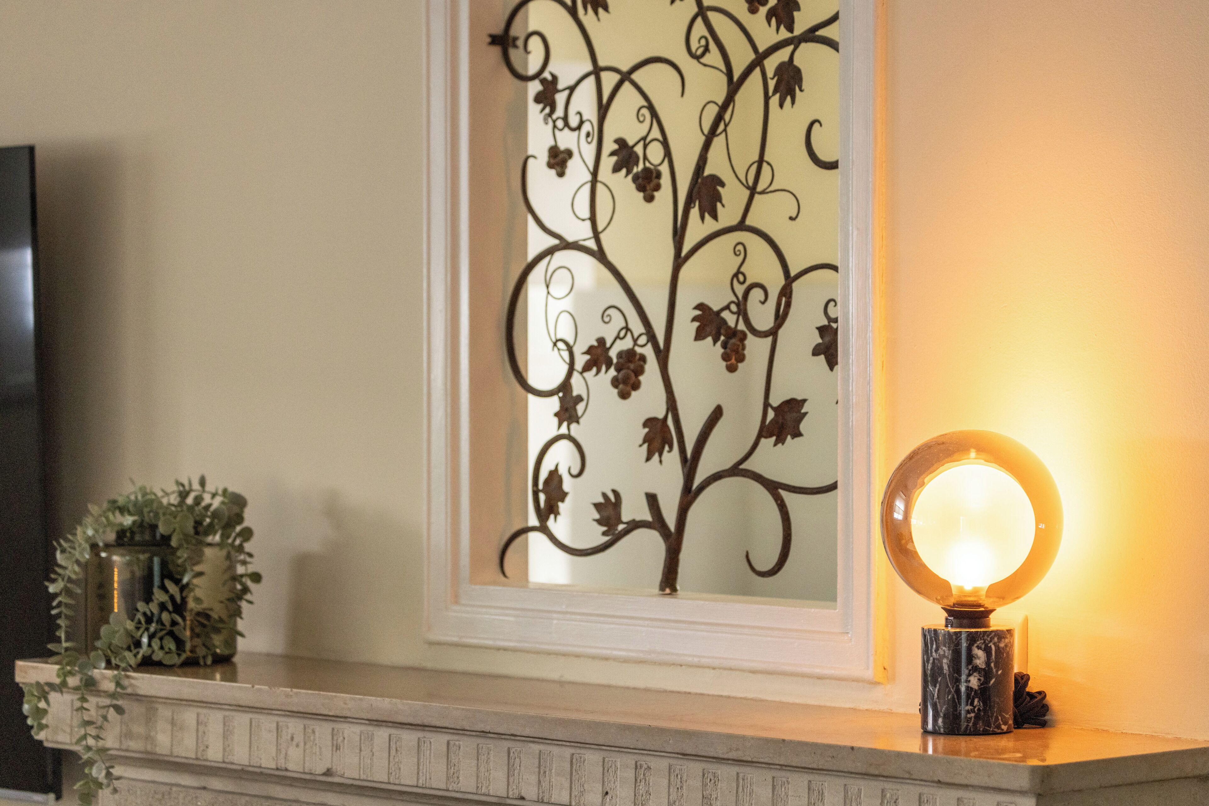 Un diseño de planta de metal decorativo en una pared, una lámpara redonda con espejo y una pequeña planta en maceta sobre una repisa.