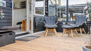 A wooden deck with two grey chairs and a small table, a glass door entrance, and a potted plant.