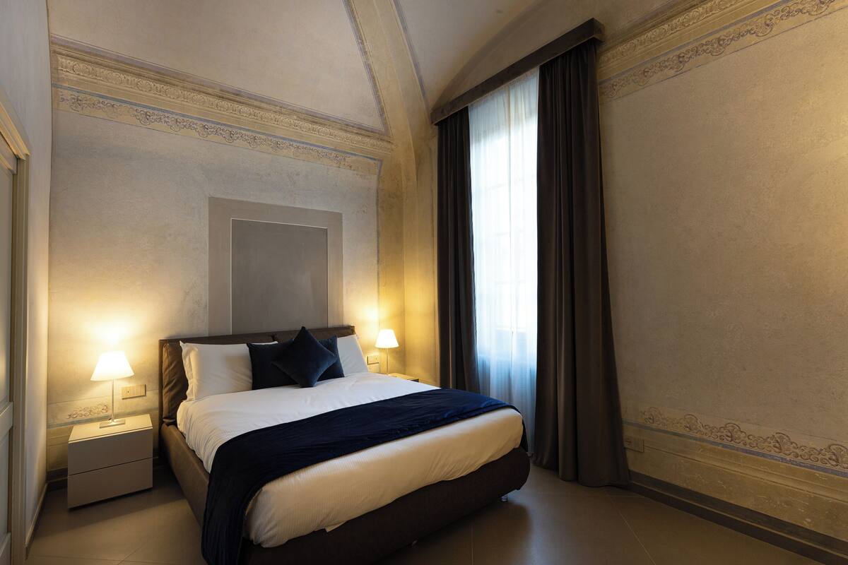 A hotel room with a large bed, two bedside lamps, a nightstand, and a window with curtains.