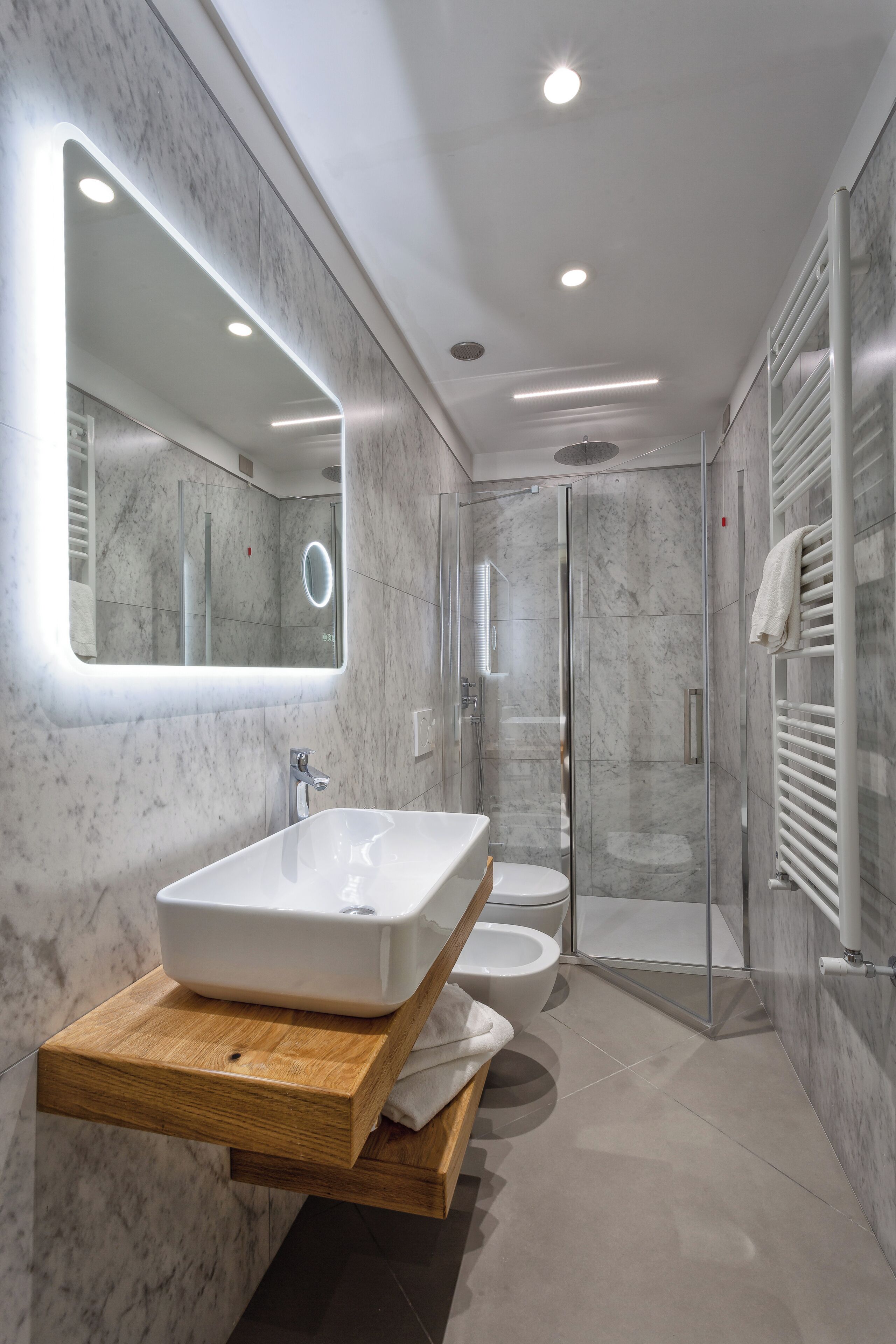 A modern bathroom with a large mirror, a wooden vanity, a glass shower enclosure, and a marble-tiled floor.