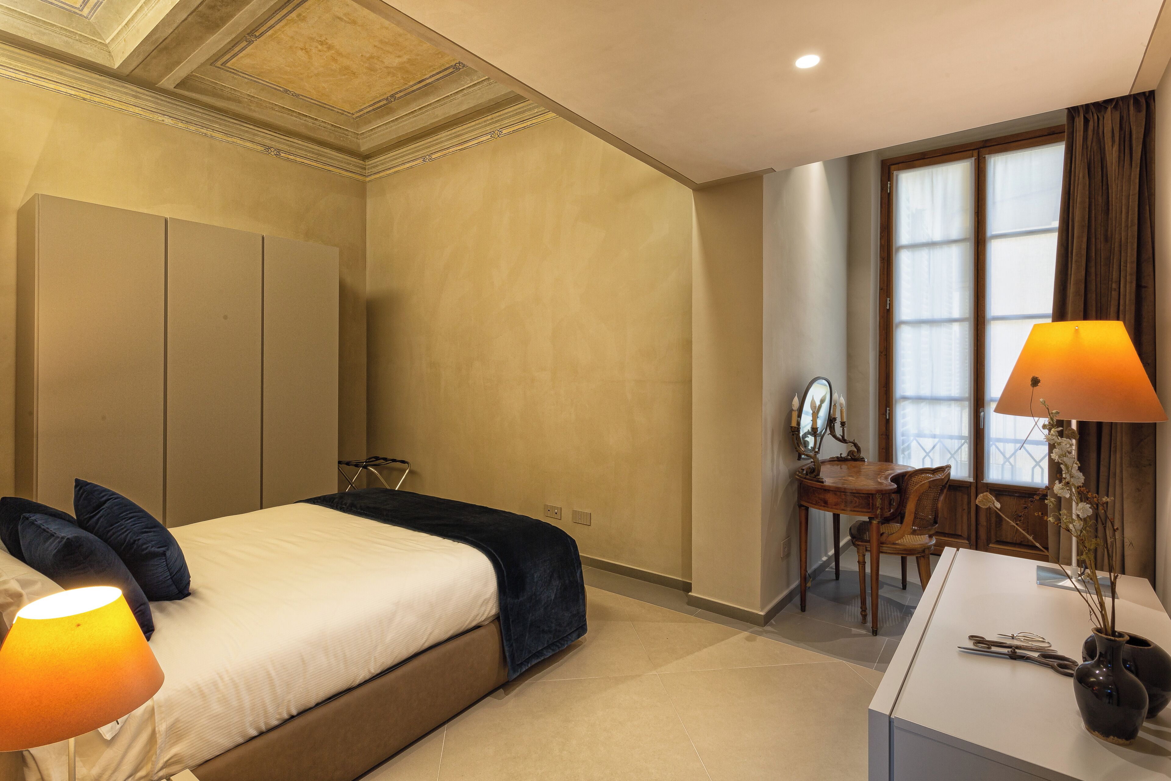 A hotel room with a bed, bedside lamps, a desk with a chair, and a window with curtains.