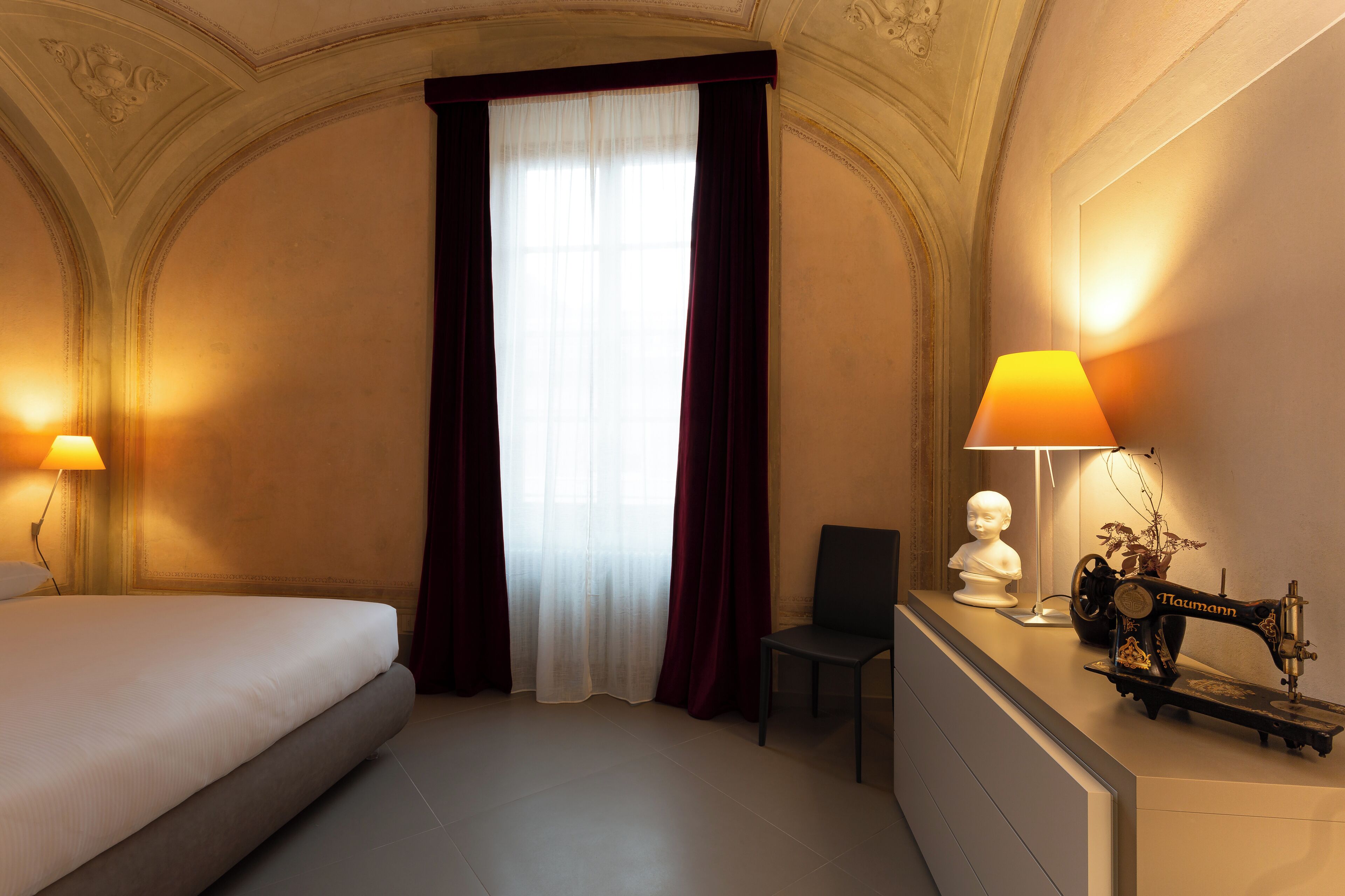 A hotel room with a bed, a chair, a lamp, a sewing machine, and a bust sculpture.