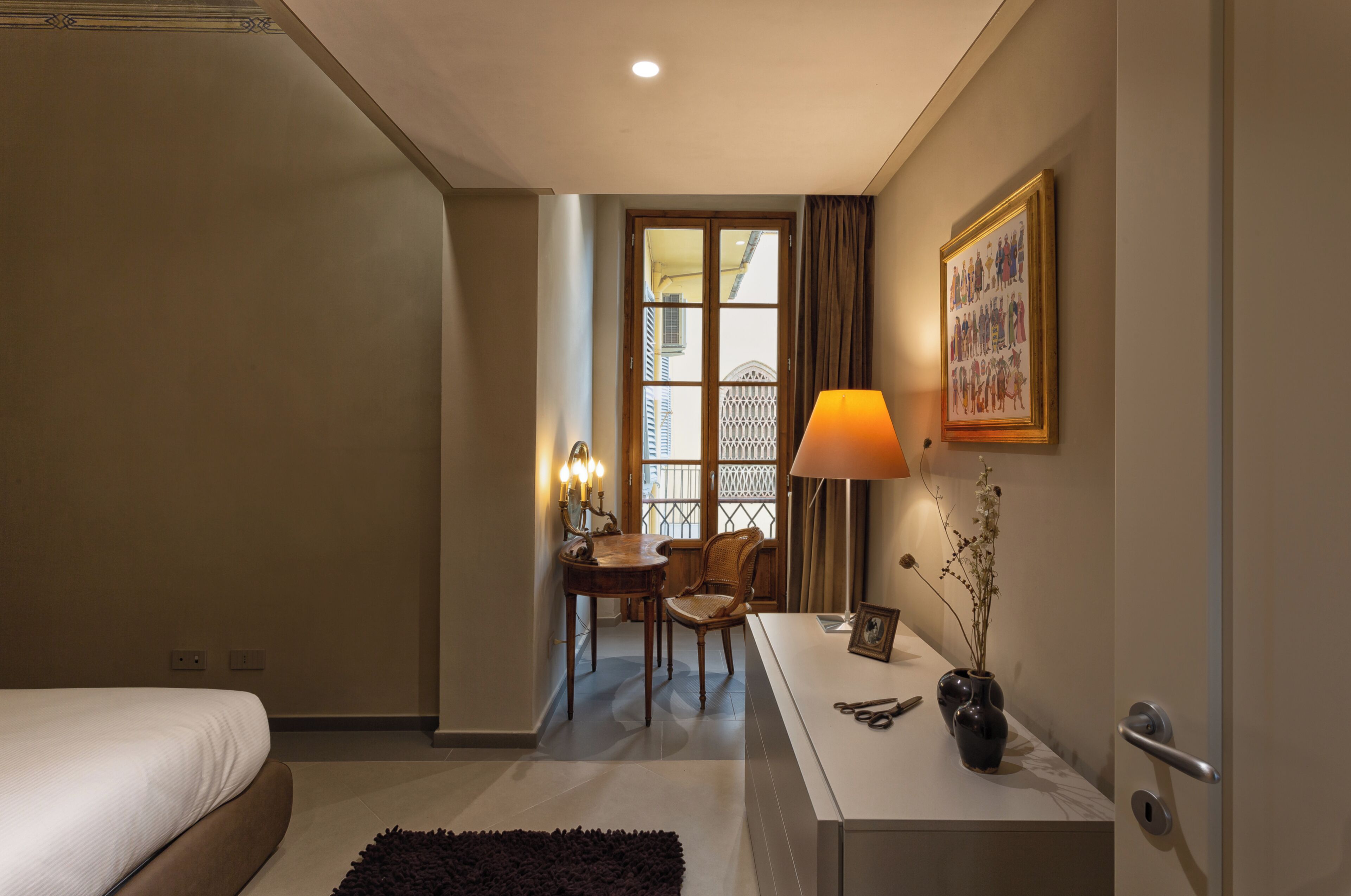 Una habitación de hotel con cama, escritorio con lámpara, silla y un jarrón con flores.
