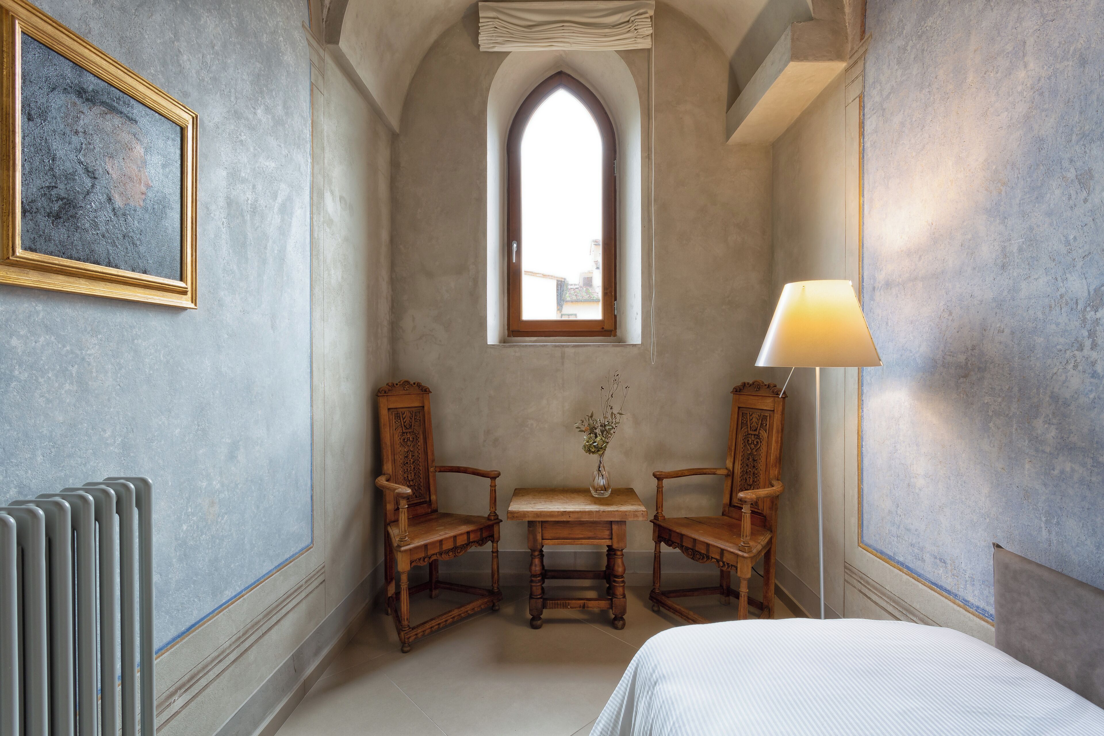 A room with a bed, a table with chairs, a window, a radiator, and a lamp.