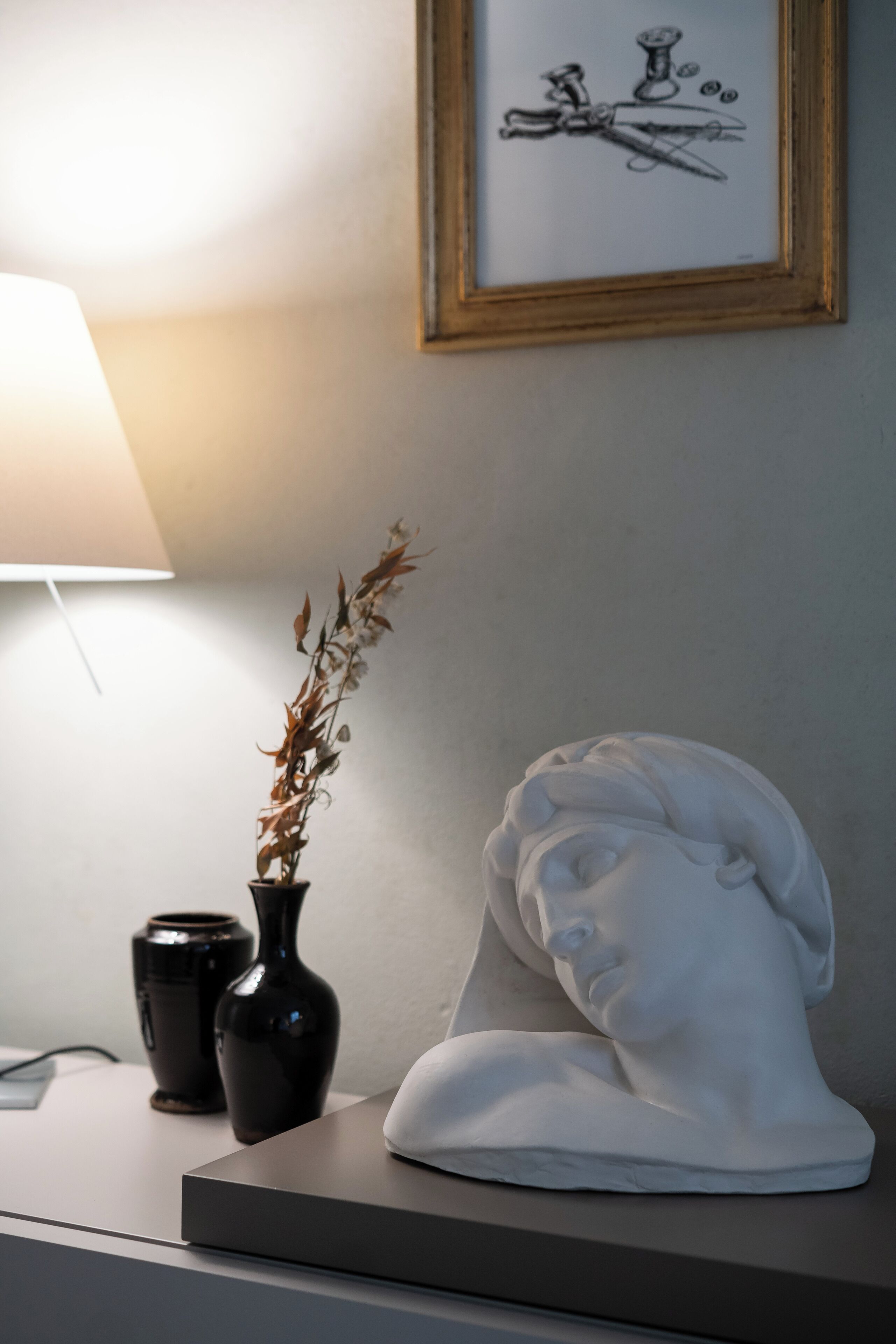 A bedside table with a vase of dried plants, a black vase, and a white bust sculpture.