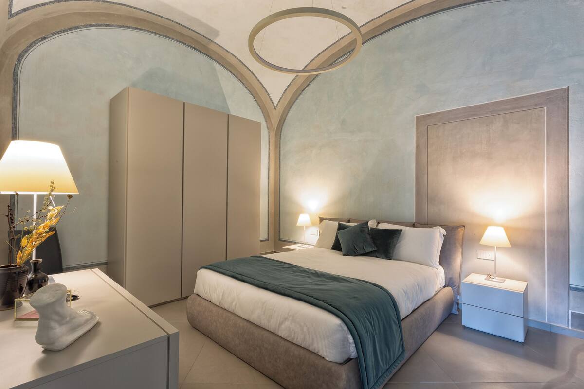 A modern bedroom with a large bed, two bedside tables with lamps, a nightstand, a dresser, and a decorative vase.