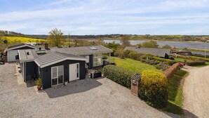 Una casa moderna con entrada de grava, un pequeño jardín y vista a un lago y campos.