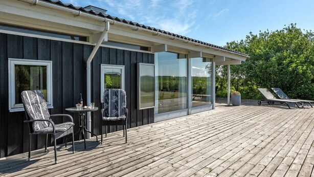 A wooden deck with two chairs and a table, a covered area with a glass wall, and a view of the surrounding greenery. - "Anund" - 1.4km from the sea by Interhome (Ringkøbing)