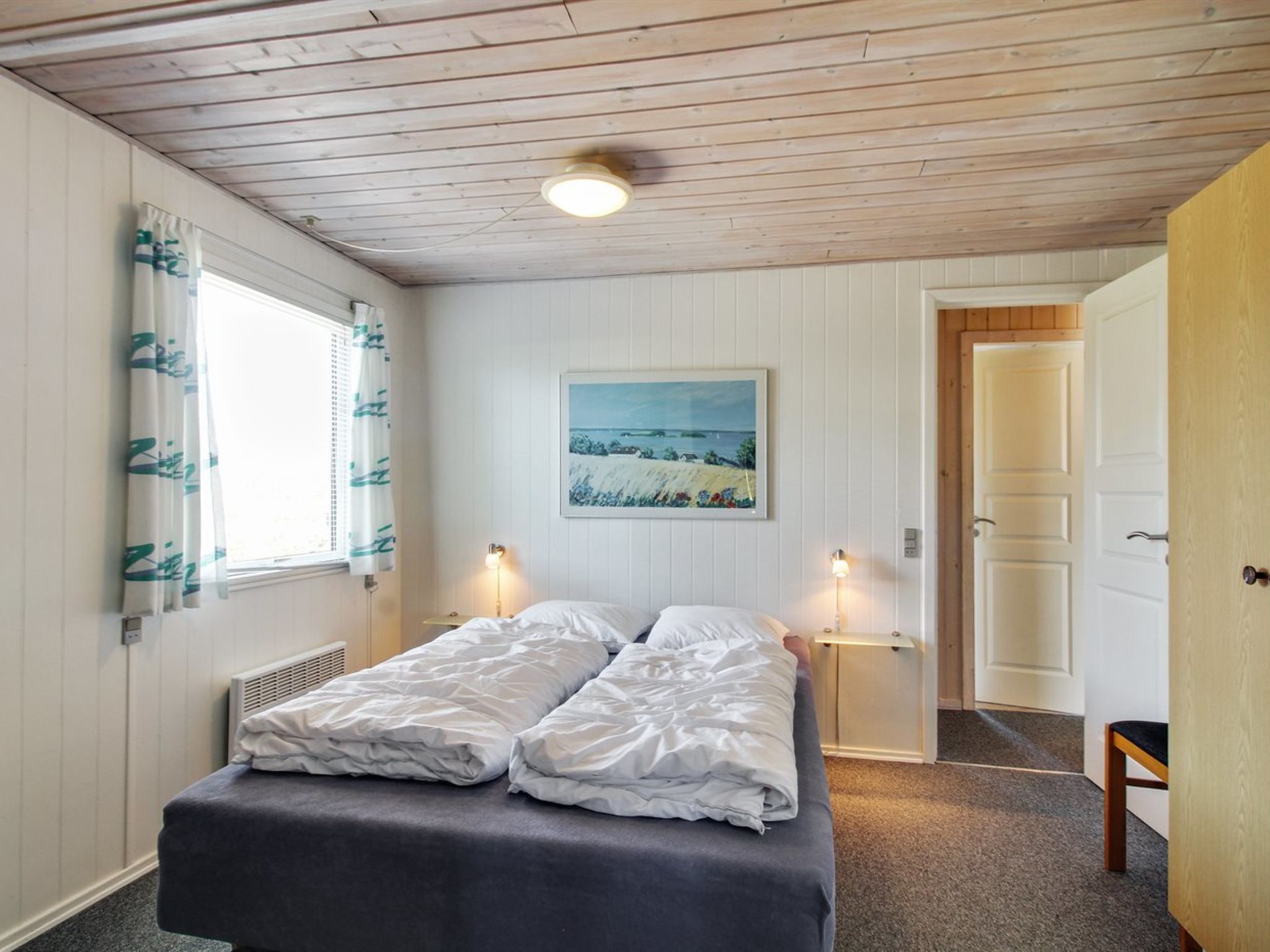 Een slaapkamer met een bed, twee bedlampen, een raam met gordijnen, een radiator, een deur en een schilderij aan de muur.