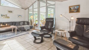 A spacious living room with a high ceiling, a large window, a sofa, two leather chairs, a coffee table, and a small side table.