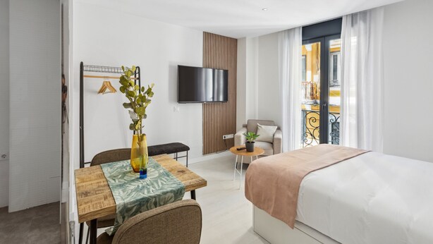 A hotel room with a bed, a desk with a plant, a chair, a TV mounted on the wall, and a balcony with a railing. - Limehome Madrid Calle de la Paloma (Madrid)