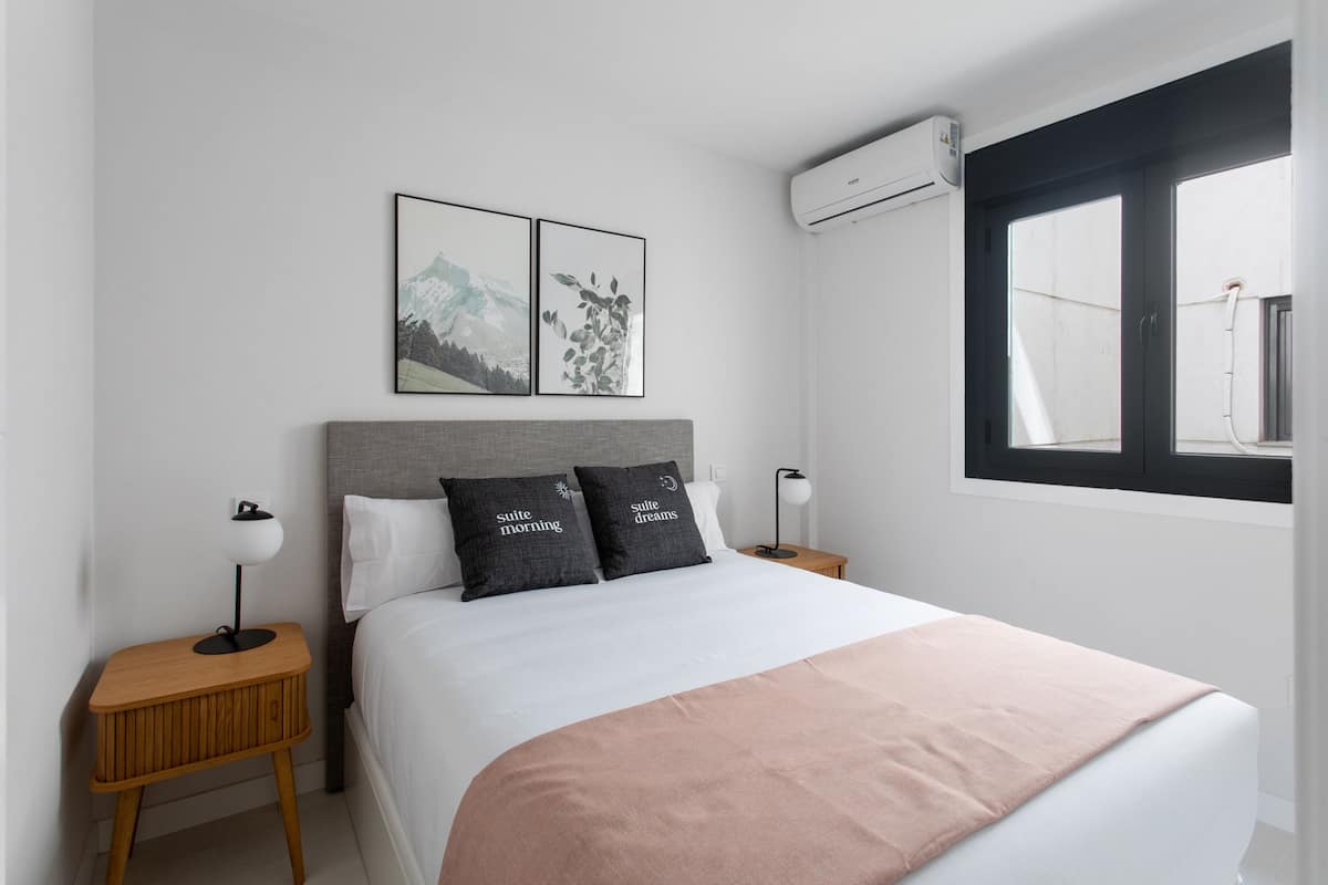 Una moderna camera d'albergo con un letto rifatto, due comodini con lampade, un'ampia finestra con vista e due quadri appesi al muro.