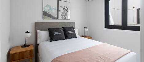 Quarto de hotel moderno com cama bem arrumada, duas mesas de cabeceira com abajures, uma janela grande com vista e duas fotos emolduradas na parede.