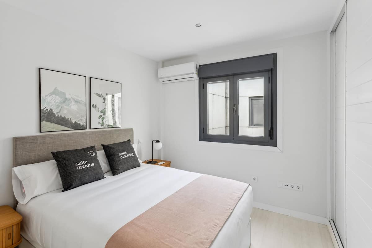 Camera da letto con un letto grande, due cuscini con scritte, un comodino, una lampada, una finestra e un condizionatore.