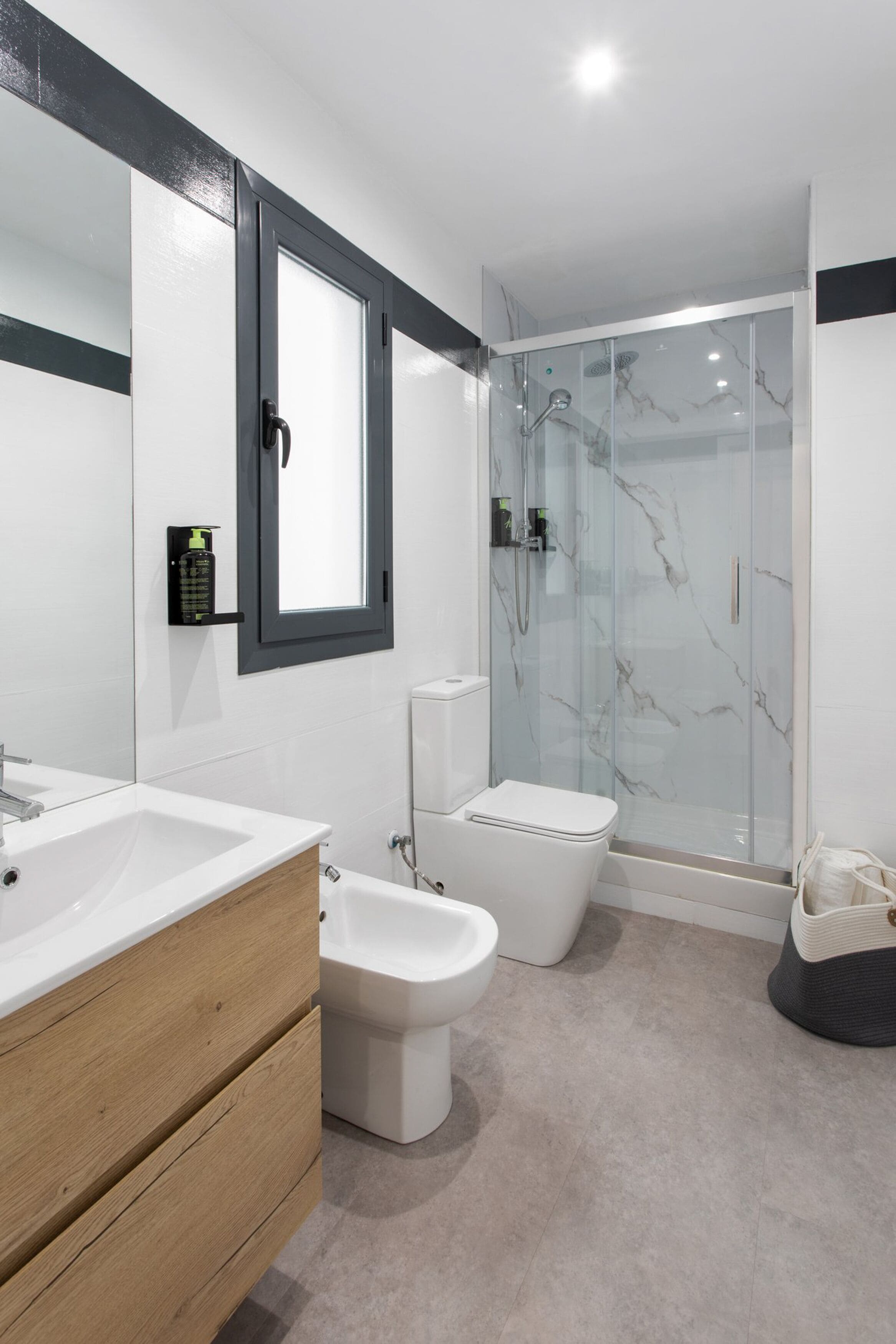Un baño moderno con un lavamanos blanco, tocador de madera, un bidé, un inodoro y un box de ducha con mampara de vidrio.