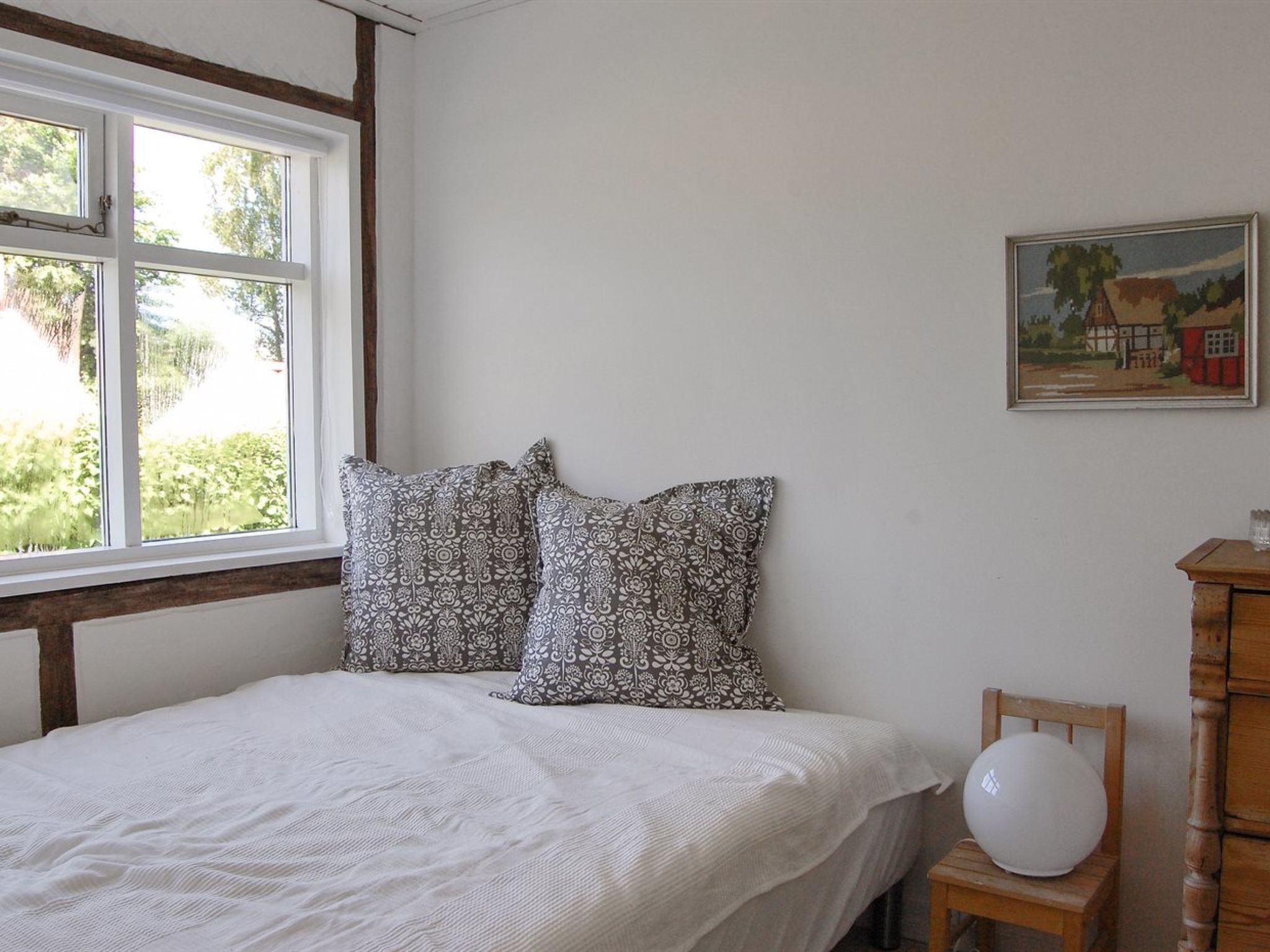 Ein Schlafzimmer mit einem Bett, zwei gemusterten Kissen, einer weißen Decke, einem Fenster, einem Holzhocker, einem kleinen Tisch und einem Bild an der Wand.