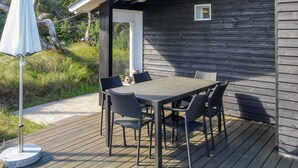 Een houten terras met een tafel en stoelen, een witte parasol en een klein raam op een donker houten gebouw.
