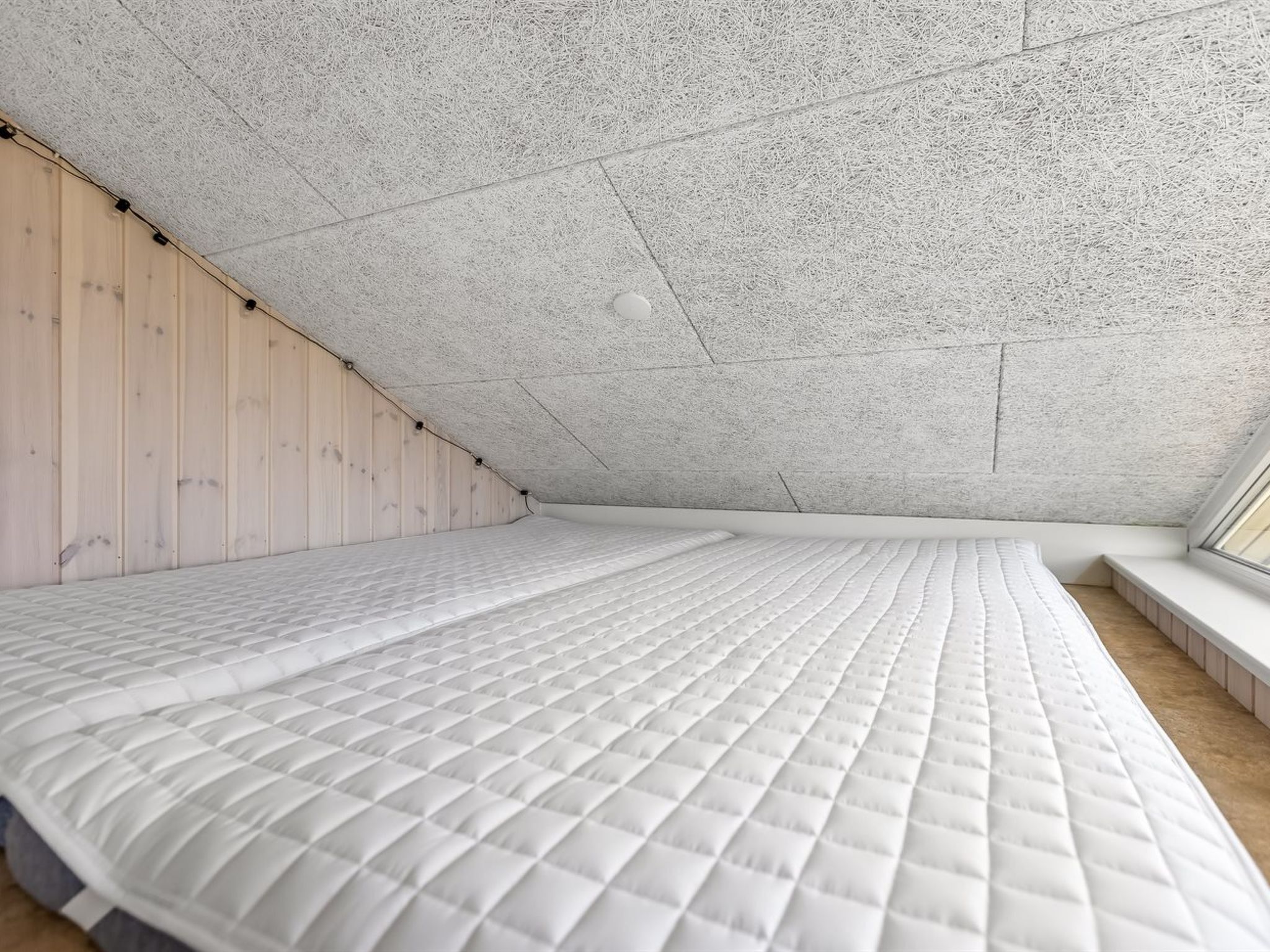 Une chambre avec un plafond en pente, un lit doté d’un matelas matelassé, un mur recouvert de lambris en bois et une fenêtre.