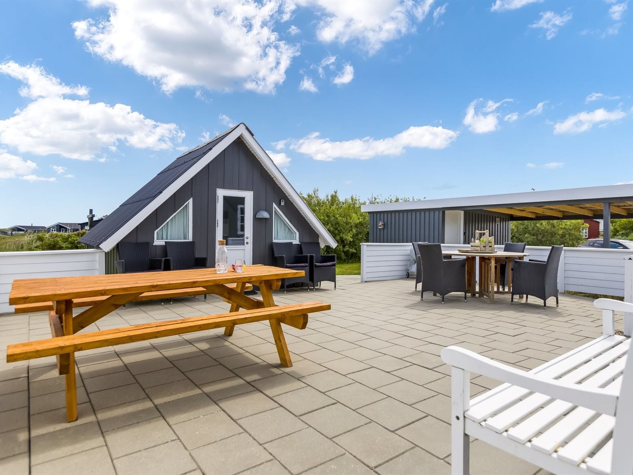 Een modern terras buiten, met een houten picknicktafel en stoelen, een overdekte zithoek en een gebouw met een schuin dak.