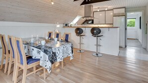 On aperçoit un coin repas avec une table dressée pour quatre personnes, des chaises en bois et deux tabourets de bar. Au fond, on distingue une cuisine équipée d’un four à micro-ondes et d’un évier.