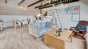 Un loft spacieux avec un plafond haut, des poutres apparentes, un coin repas avec une table et des chaises, un canapé, une table basse avec des boissons et un escalier menant à un niveau supérieur.
