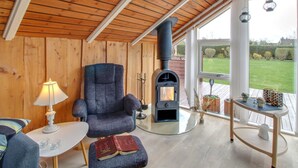 Ein gemütliches Zimmer mit Holzbalkendecke, einem bequemen Sessel, einem Tisch mit Lampe, einem Buch und einem modernen Kamin.