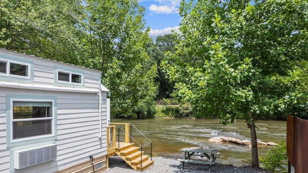 A white RV with a gravel patio, a picnic table, and a wooden staircase leading to a deck overlooking a river.