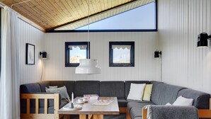 Una sala de estar moderna con techo de madera, un sofá seccional gris, una mesa de centro de madera y dos ventanas con marcos negros.