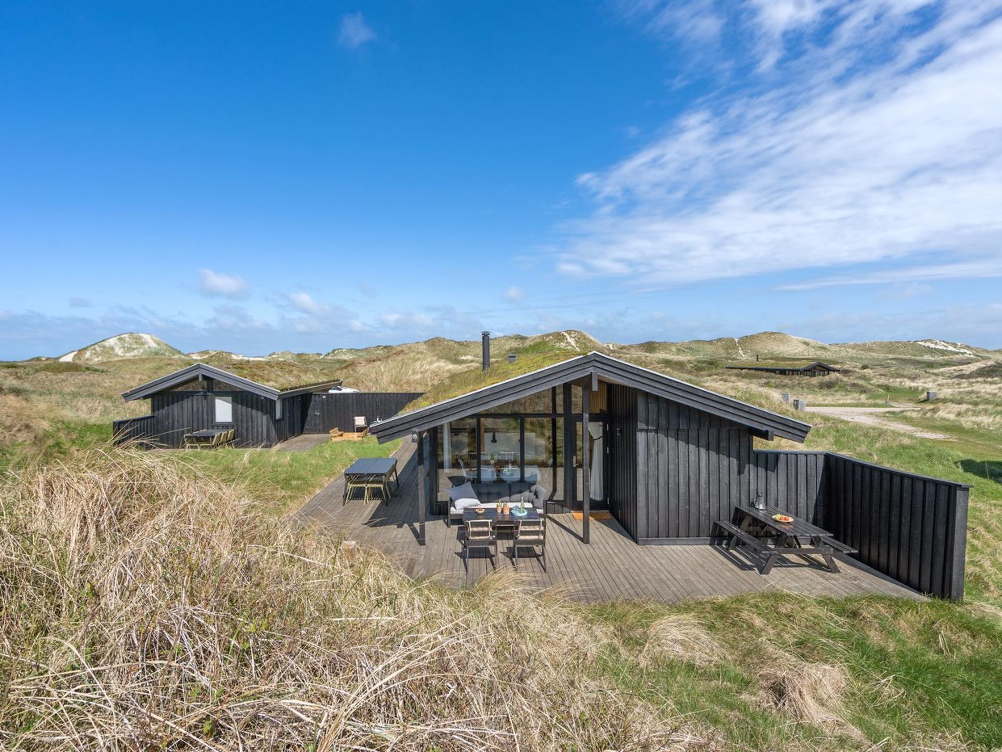 A modern wooden cabin with a deck, large windows, and a view of the dunes.