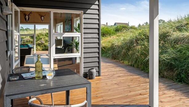 Un pontile in legno con tavolo e sedie, una porta vetrata che conduce a una stanza con tavolo da biliardo e vista sul paesaggio circostante.